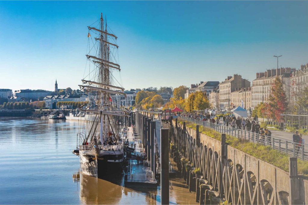 Le Belem accosté sur le quai de Nantes. Une partie de la ville de Nantes et la promenade longeant la Loire sont visible autour du bateau.