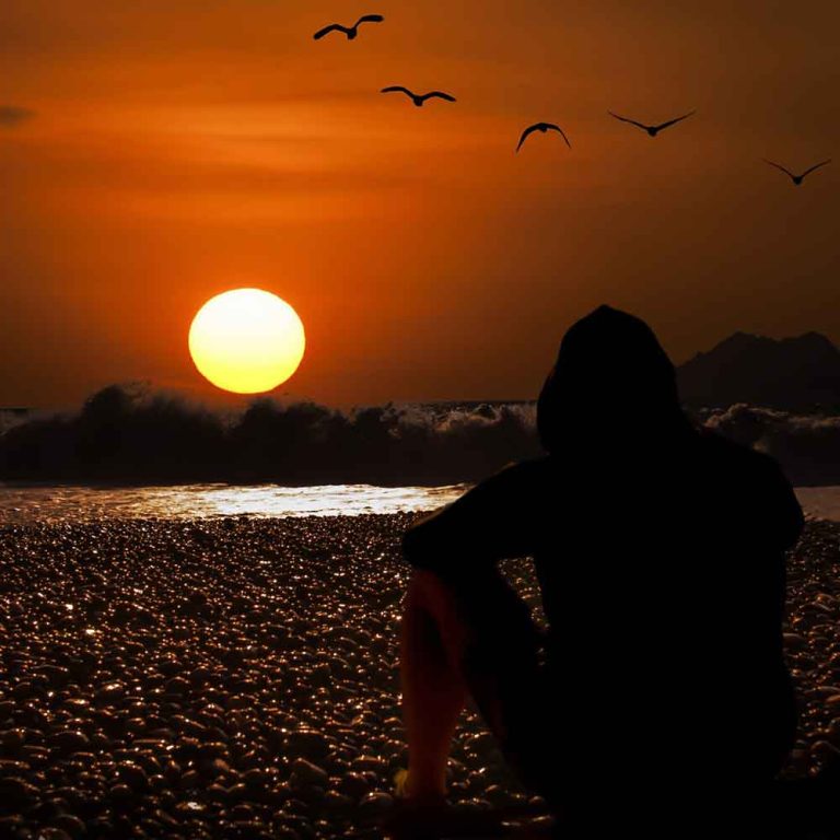 Une personne assise sur une plage de galets au coucher de soleil. Le ciel est orangé et une vague arrive.